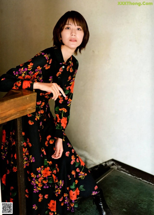 A woman in a black floral dress leaning against a wall.