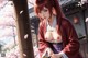 A woman in traditional attire stands near a wooden pillar with cherry blossoms and a building in the background.